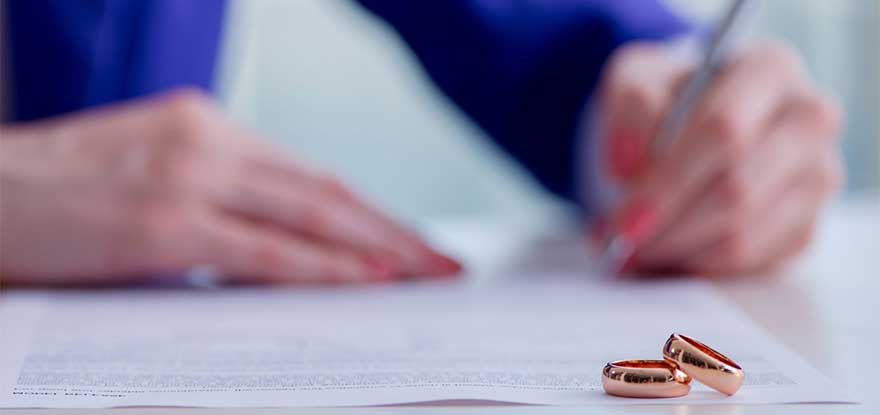 Two wedding rings sit on a table in the foreground while a person with red-painted nails, possibly advised by Tinley Park family law attorneys, signs a document in the background.